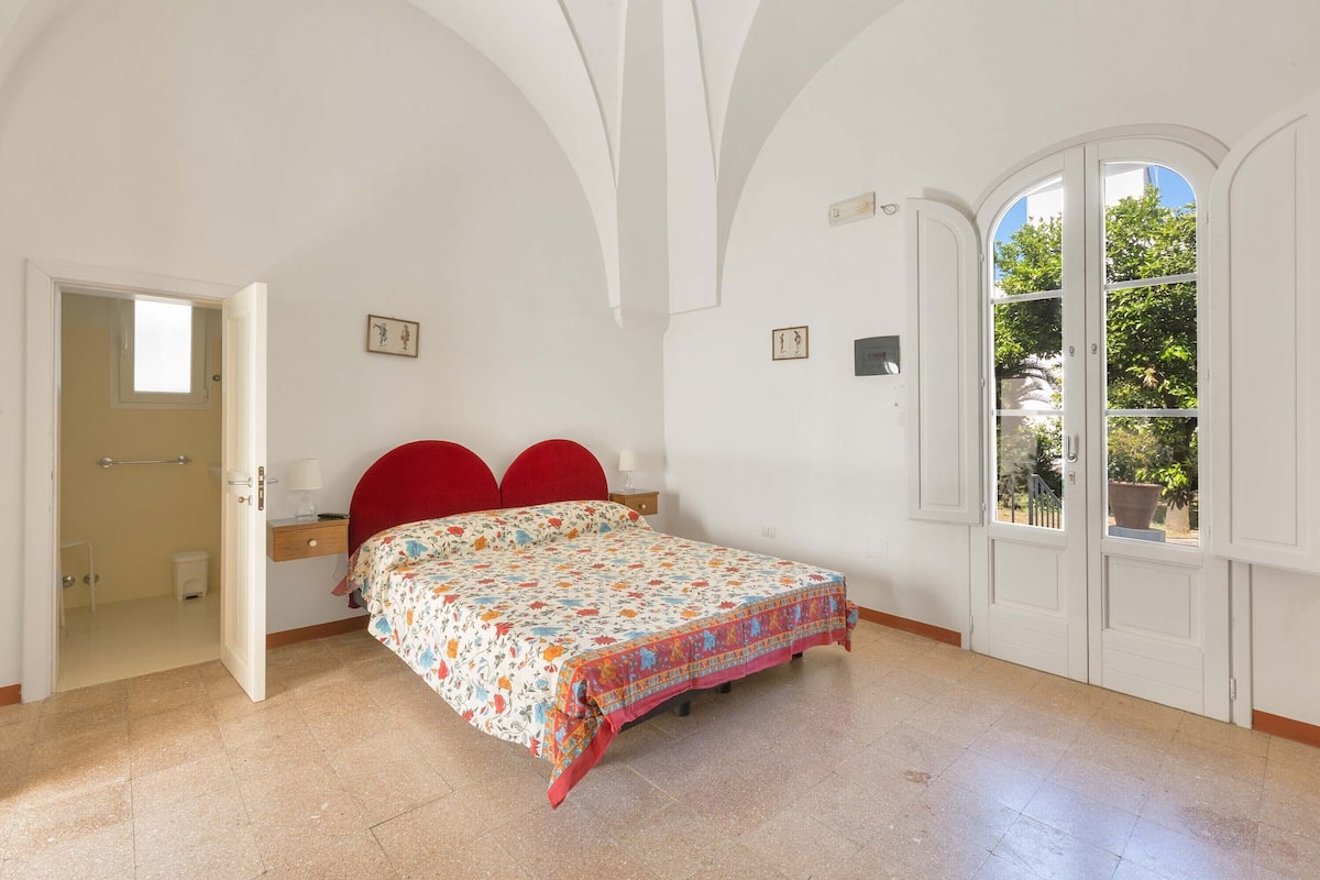 A bedroom with a bed, a red headboard, a small bedside table with a lamp, and a window with a view of greenery.