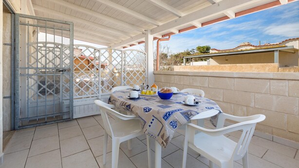 A covered outdoor patio with a table set for two, white plastic chairs, and a view of a building in the background. - Residence Aquarium (Torricella)