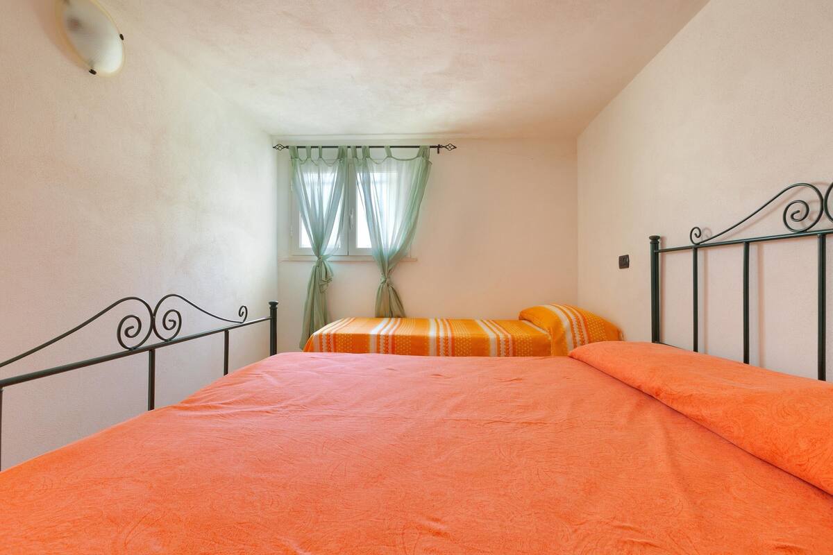 A single bed with an orange bedspread, a window with sheer curtains, and a wall-mounted light fixture.