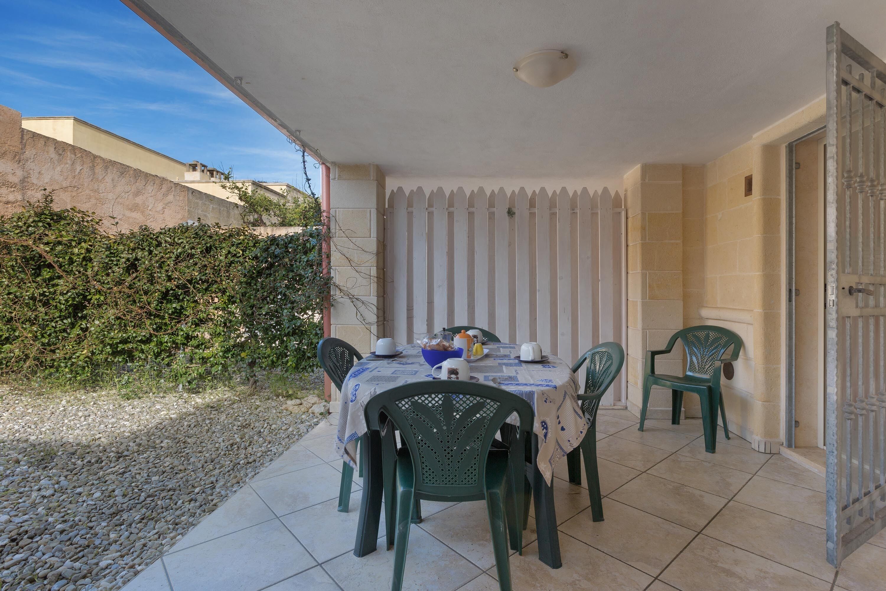 An outdoor patio with a table set for four, surrounded by green chairs, a white tablecloth, and a white fence.