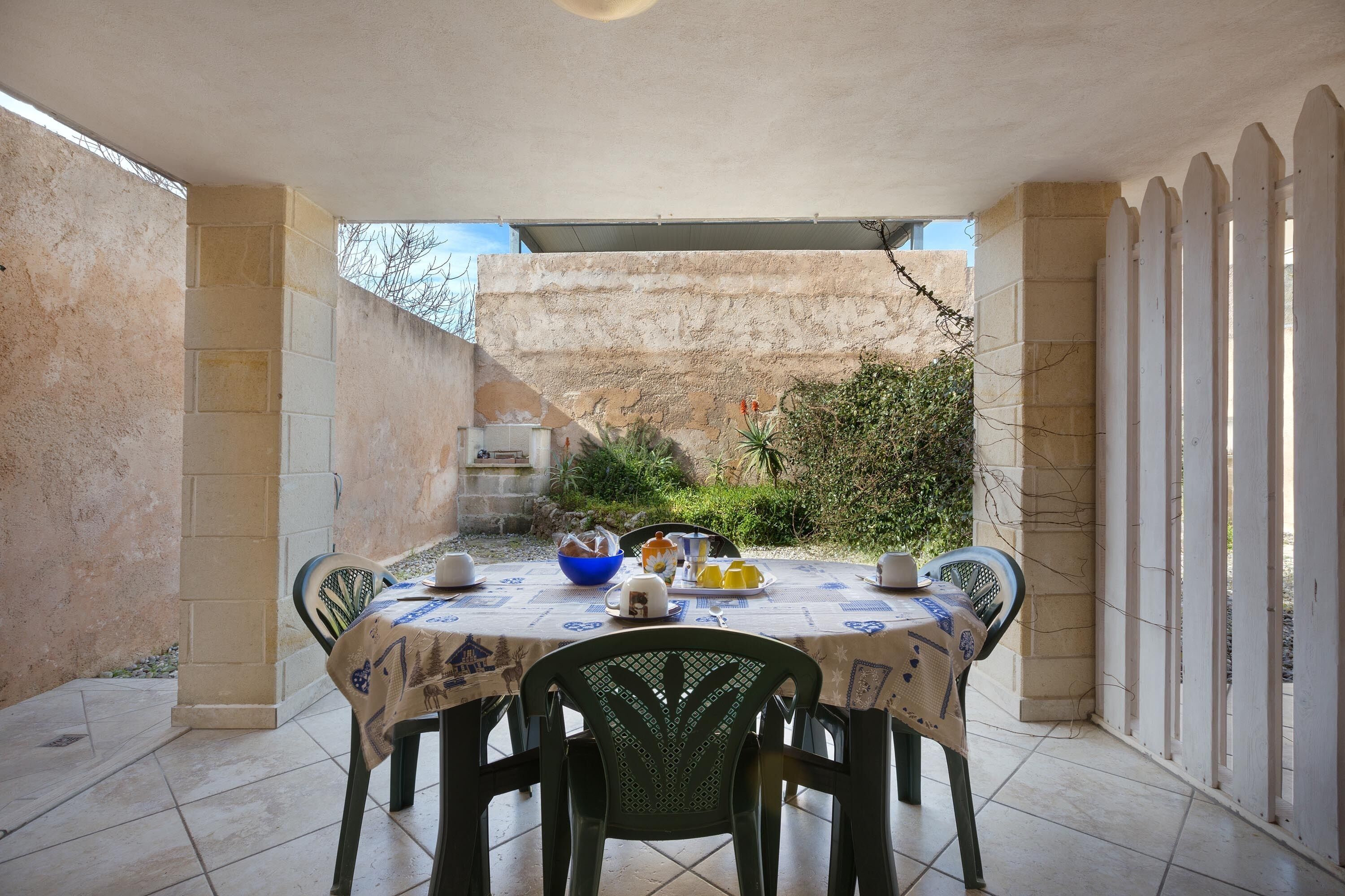 An outdoor patio with a round table set for dining, surrounded by chairs, and a white picket fence.