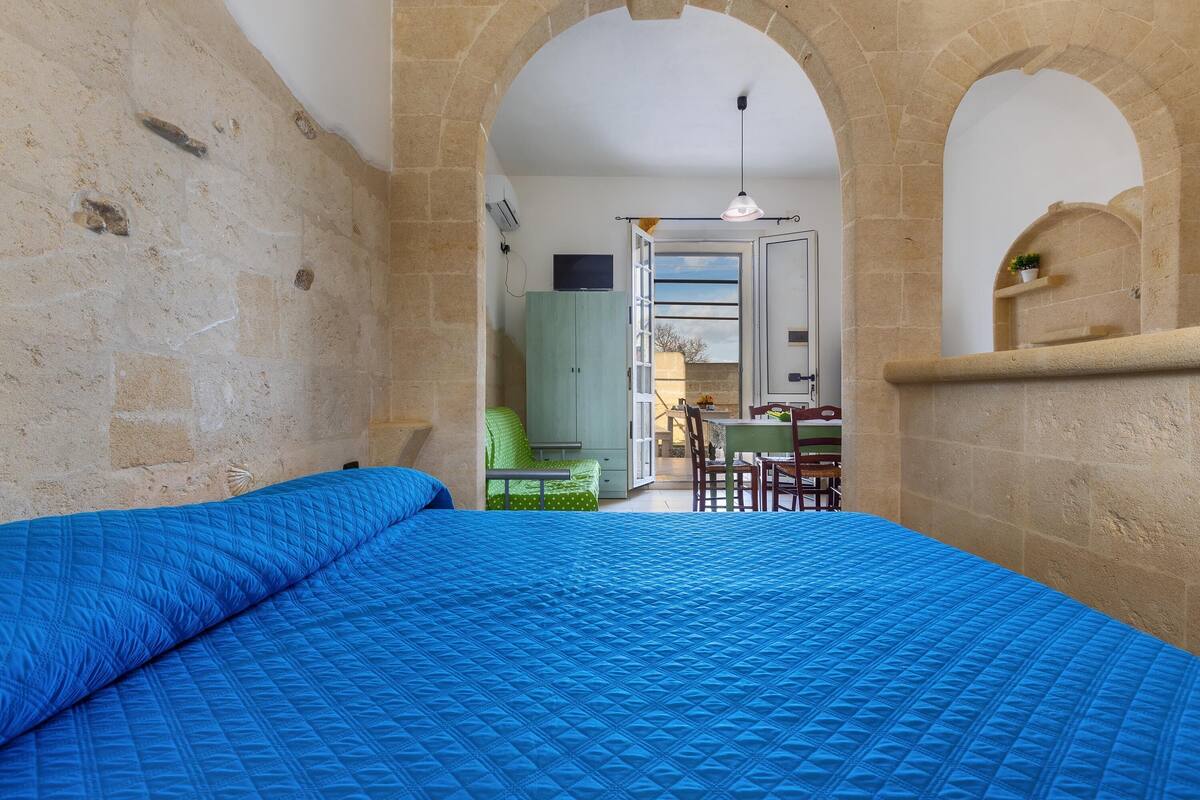 A room with a bed, a desk, and a chair. There is a window with an open door leading to another room.