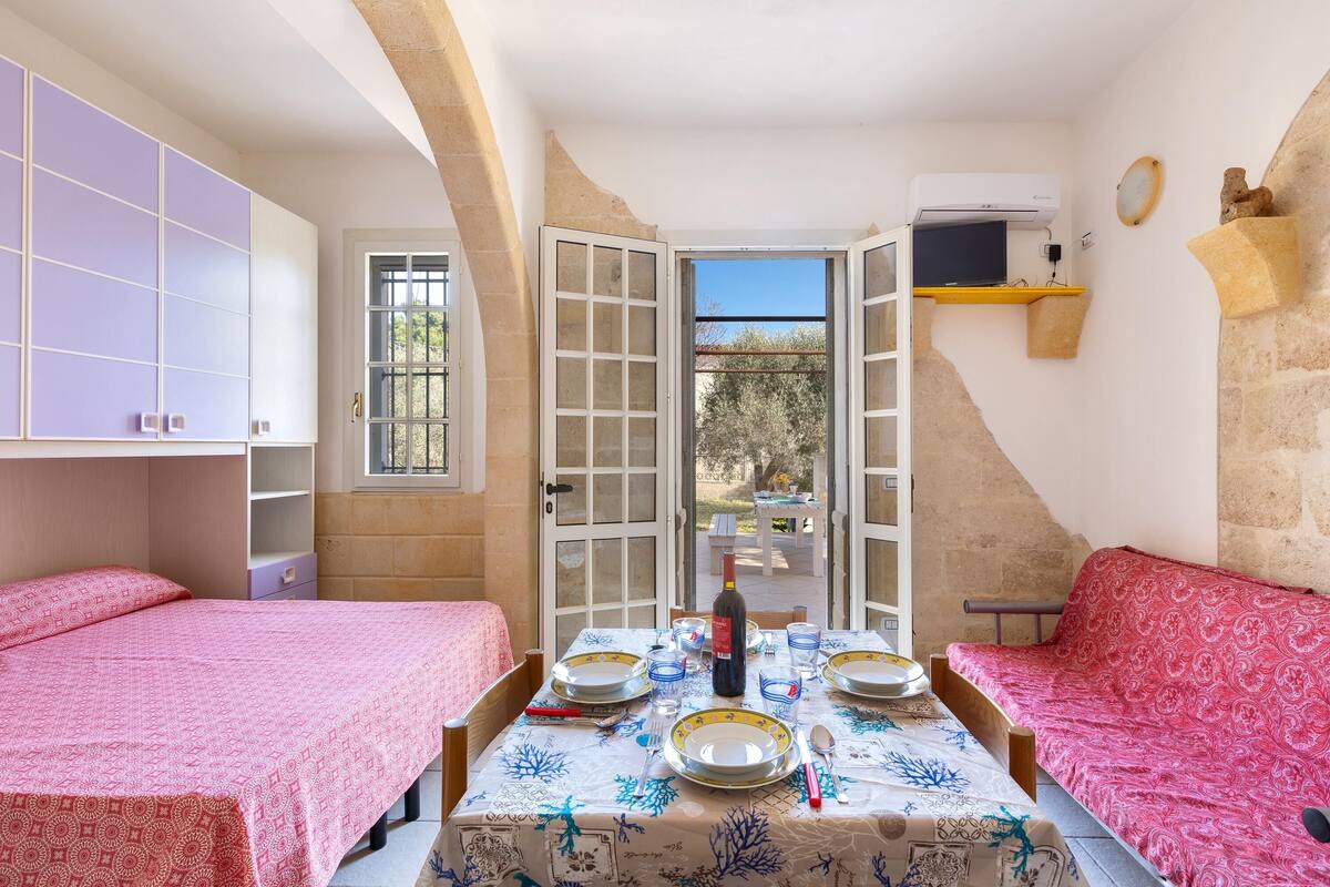 A cozy room with two beds, a dining table set for four, and a view of the garden through open doors.