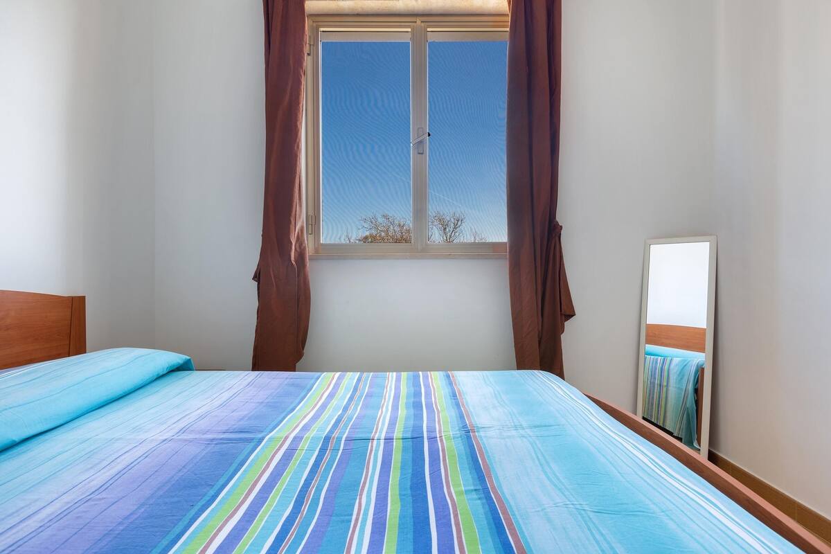 A bedroom with a large window, a bed with striped bedding, and a mirror on the wall.