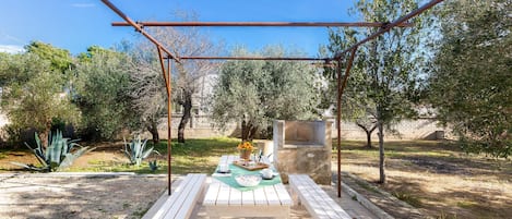 An outdoor patio with a table set for two, surrounded by olive trees and a pergola structure.