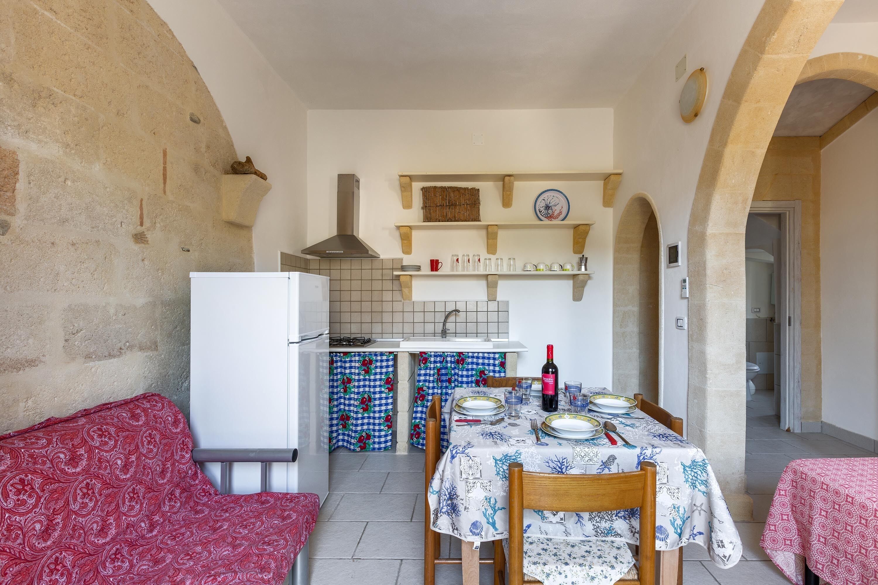 A compact kitchen with a refrigerator, sink, and wooden shelves. A dining table with plates and a wine bottle is set in the dining area.