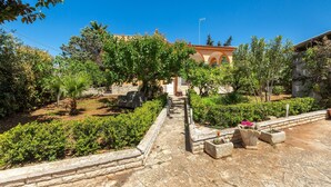 Une cour avec un chemin de pierre menant à un bâtiment orné d’arcades, entouré de verdure et de plantes en pot.