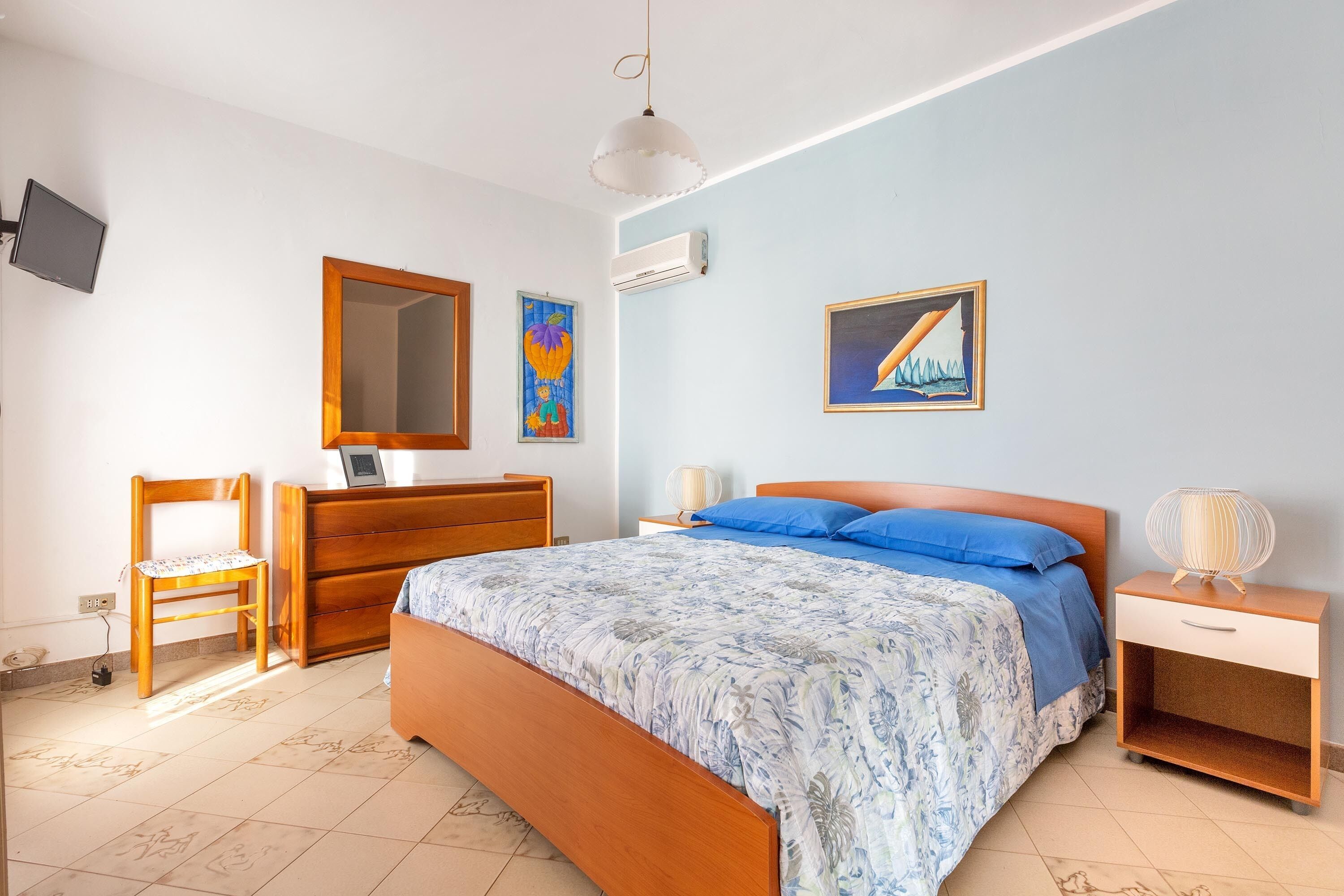 A bedroom with a bed, a wooden dresser, a chair, a bedside table, and a wall-mounted TV.