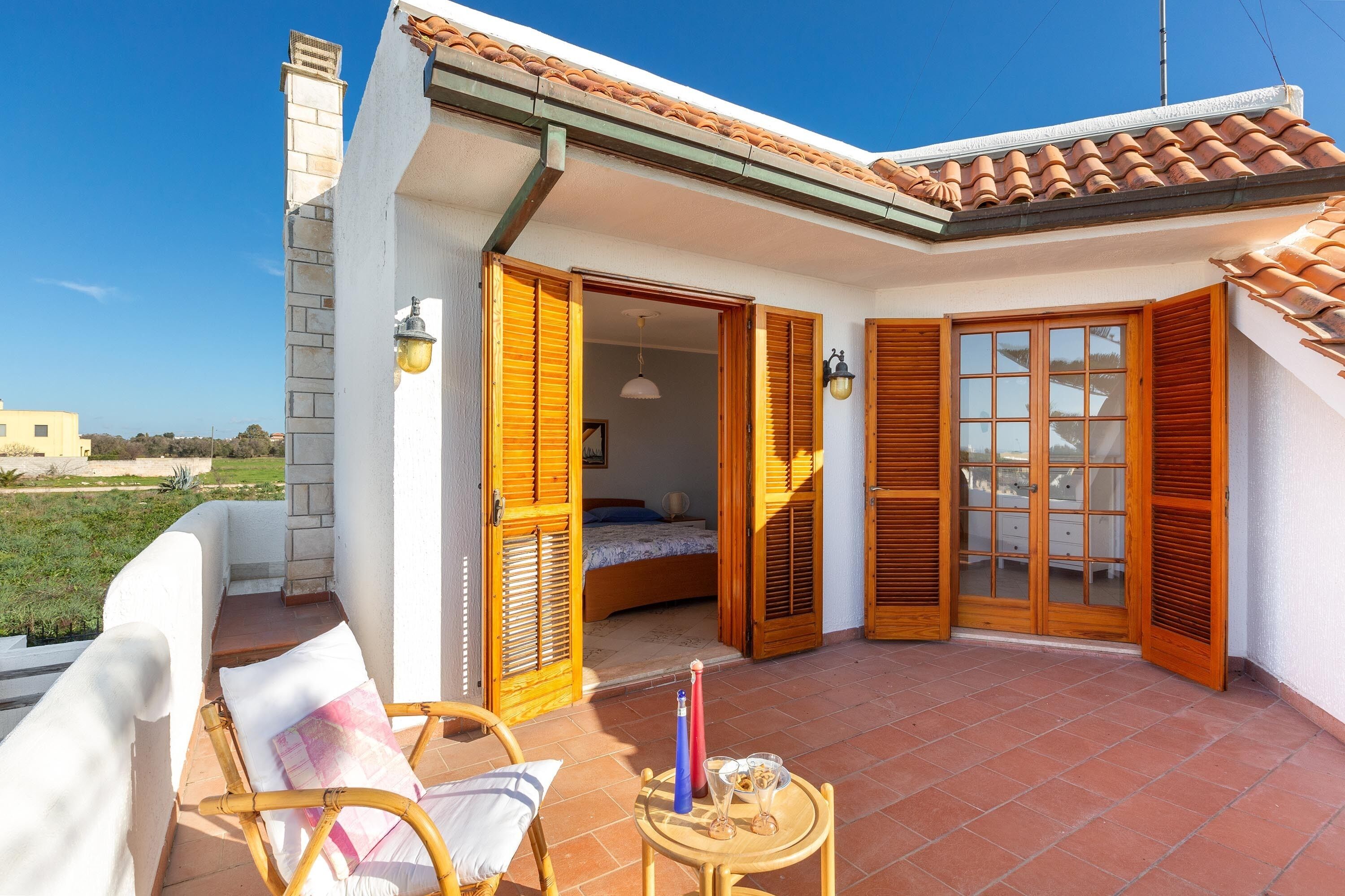A small, white house with a tiled roof, wooden shutters, and a balcony with wicker furniture.