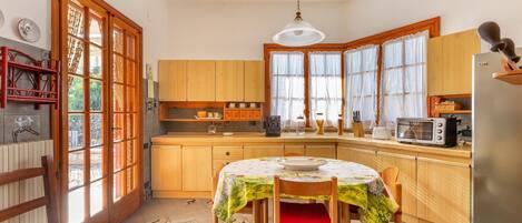 A kitchen with wooden cabinets, a dining table with a tablecloth, a refrigerator, and a window with curtains.