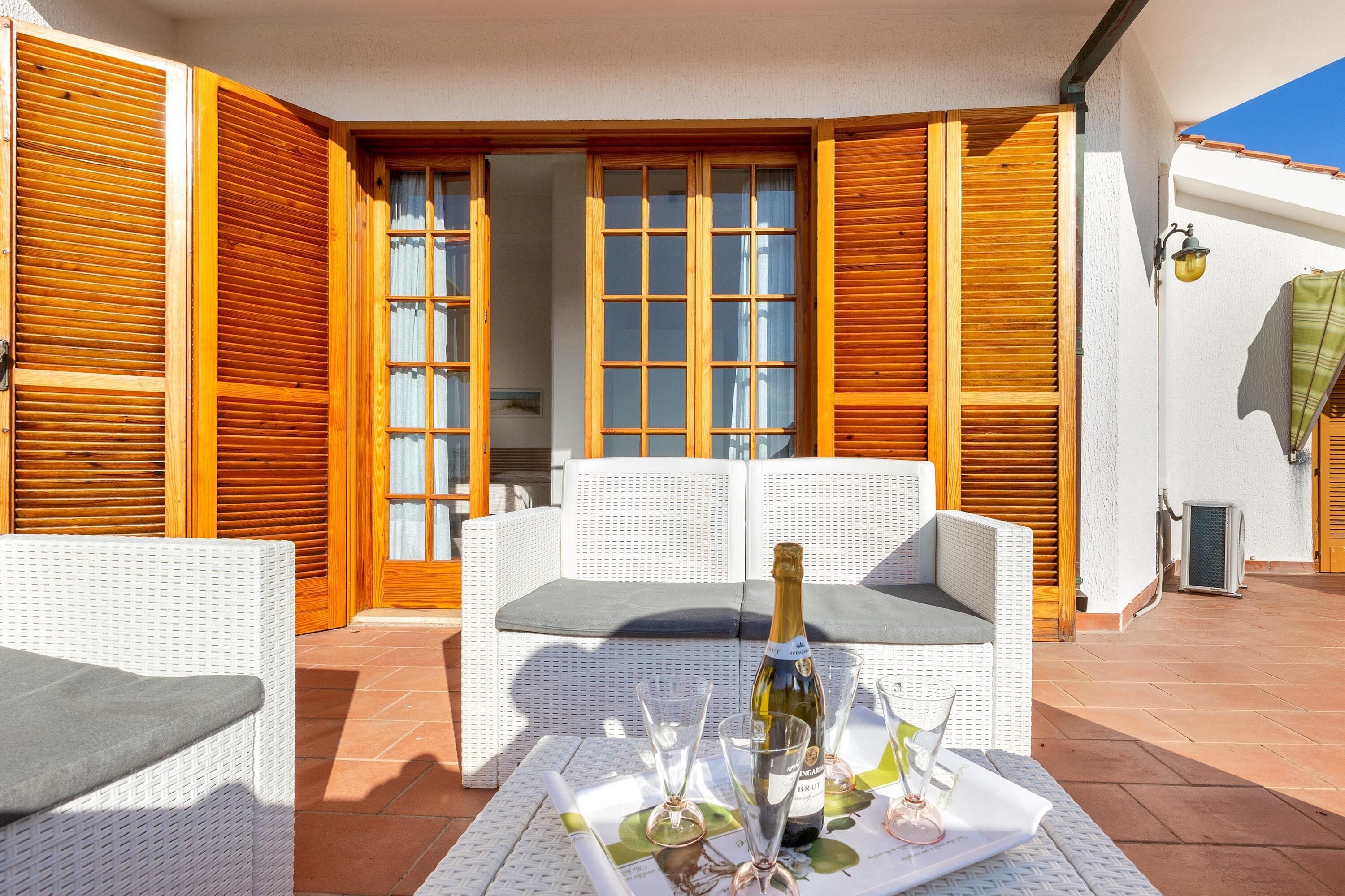 Un patio con un sofá blanco de mimbre, una mesa con una botella y copas, y contraventanas de madera.