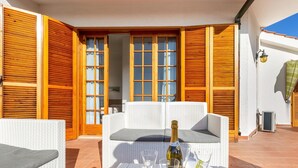 A patio with a white wicker sofa, a table with a bottle and glasses, and wooden shutters. - Villa Alice (Porto Cesareo)