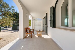 A covered outdoor area with a bench and table, white walls, and a view of a gate and trees.