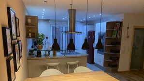 A modern kitchen with a central island, pendant lights, and a dining area with a wooden table and white chairs. - Beautiful and cozy house in the middle of nature. (Niedereschach)