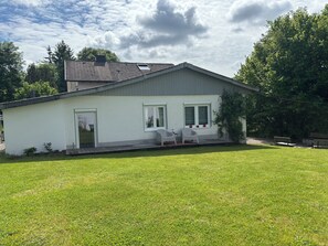Uma casa térrea com fachada branca, telhado cinza e um pequeno alpendre com duas cadeiras brancas.