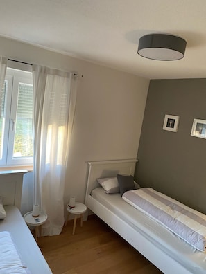 Un dormitorio con una cama individual, una mesita de noche, un taburete pequeño y una ventana con cortinas.