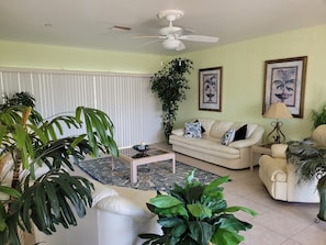 Un salon avec un canapé blanc, une table basse et diverses plantes. On y trouve également un ventilateur de plafond et une fenêtre avec des stores.