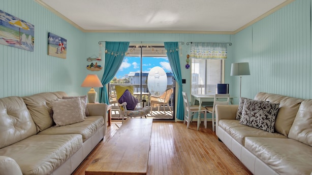 A living room with two beige sofas, a wooden coffee table, a small white table, and a chair. There are curtains, a lamp, and a window with a view of the sky. - Savannah Beach & Racquet Club 303A (Tybee Island)