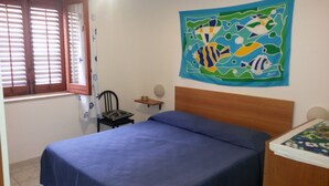Un dormitorio con cama, una silla, una mesita, una ventana con persianas y un tapiz de colores.