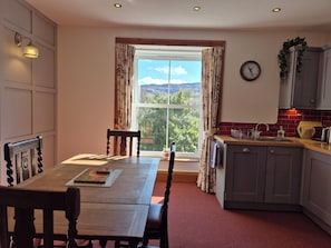 A dining area with a table and chairs, a window with curtains, a kitchen with a sink and cabinets, and a wall clock.