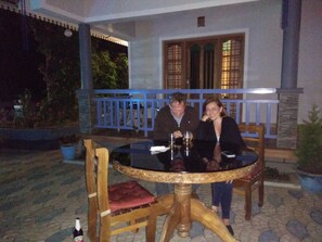 Una pareja sentada en una mesa redonda con una copa de vino, en un balcón con barandilla azul y plantas en macetas.
