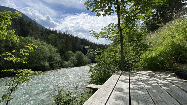 Une terrasse en bois donnant sur une rivière, avec une montagne boisée en arrière-plan.