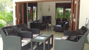 A living room with wicker furniture, a television, and a view of greenery outside. - Lombok From Your Private Villa Sepuluh (Senggigi)