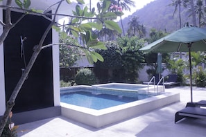 Une piscine rectangulaire bordée de blanc, entourée d’une terrasse également blanche. On y trouve des chaises longues et un parasol vert sous un palmier.