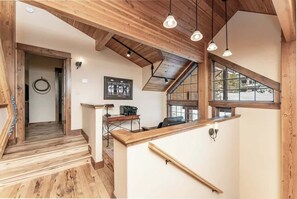 A wooden staircase leading to a loft area with a desk and chair, a large window, and a ceiling with exposed beams.