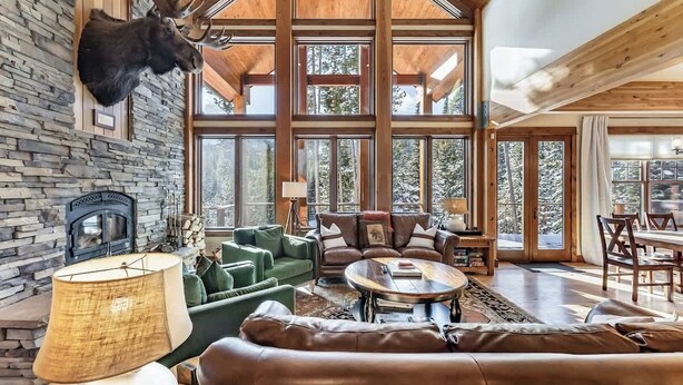 A spacious living area with a stone fireplace, leather sofas, and a large window offering a view of the outdoors. - Moose Creek | Ski-In/Out + Hot Tub (Big Sky)