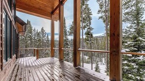A wooden deck with a railing, overlooking a forested area with snow-covered trees. - Moose Creek | Ski-In/Out + Hot Tub (Big Sky)