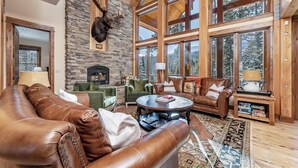 A spacious living room with a stone fireplace, leather furniture, and large windows offering a view of the outdoors. - Moose Creek | Ski-In/Out + Hot Tub (Big Sky)