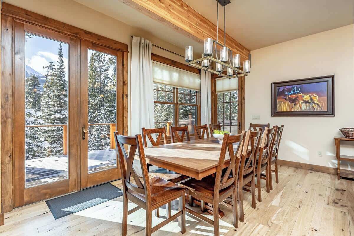 A dining area with a wooden table and chairs, large windows with a view of snow-covered trees, and a chandelier.