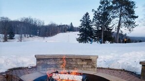 A stone fire pit with a fire burning, surrounded by a snowy landscape and trees.