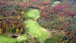 An aerial view of a golf course surrounded by dense forest with autumn foliage.