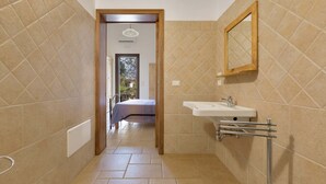Un baño estrecho con baldosas color beige, un lavamanos blanco con espejo y una puerta que da a un dormitorio.