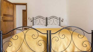 Una cama con estructura metálica y colcha amarilla en una habitación con paredes blancas y una puerta de madera.