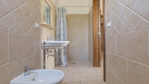 Un baño estrecho con azulejos color beige, un lavamanos blanco, un espejo y una cortina de ducha.