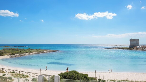 Une plage avec une eau d’un bleu limpide, un rivage sablonneux et une petite construction en forme de tour.