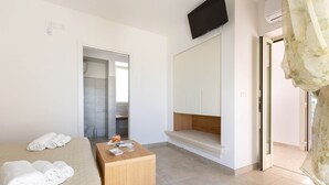 Une chambre d’hôtel avec un lit, une table de chevet en bois, un téléviseur fixé au mur et une salle de bain visible à travers une porte ouverte.