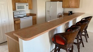 Eine Küche mit Holzausbau, einem weißen Kühlschrank, einer Mikrowelle und einem Herd. Vier Holzbarhocker mit roten Sitzkissen sind vorhanden.