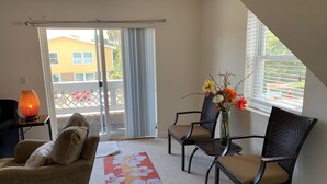 Ein Wohnzimmer mit einer Couch, zwei Sesseln, einem Couchtisch, einer Stehlampe und einer Vase mit Blumen. Eine Glastür führt auf einen Balkon mit Blick auf ein Gebäude und einen Zaun.