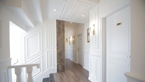 Un couloir aux murs blancs, au plancher en bois et avec un mur d’accent en marbre. On y trouve des portes avec des plaques numérotées et des tableaux encadrés accrochés au mur.
