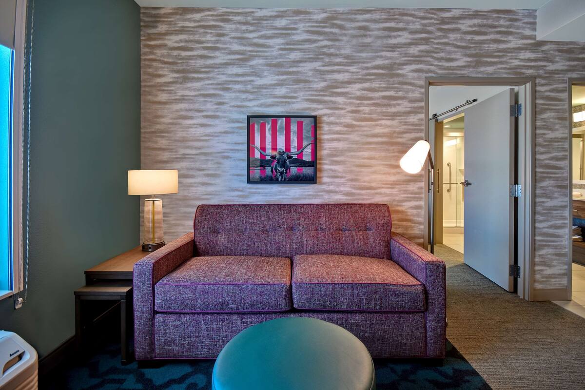 A hotel room with a purple sofa, a round ottoman, a lamp, and a painting of a bull on the wall.