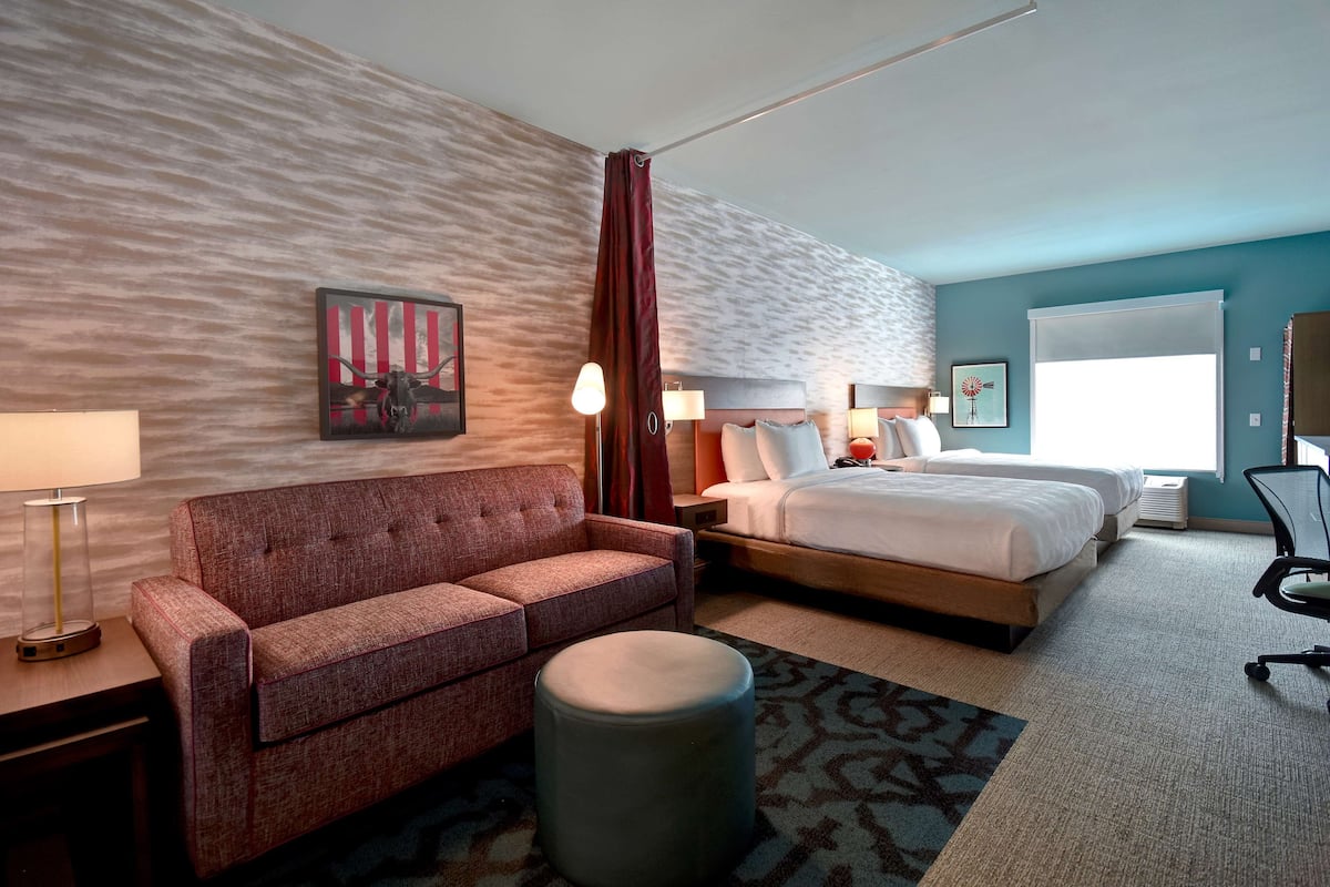 A hotel room with a bed, a sofa, a round ottoman, a desk with a lamp, and a window with curtains.