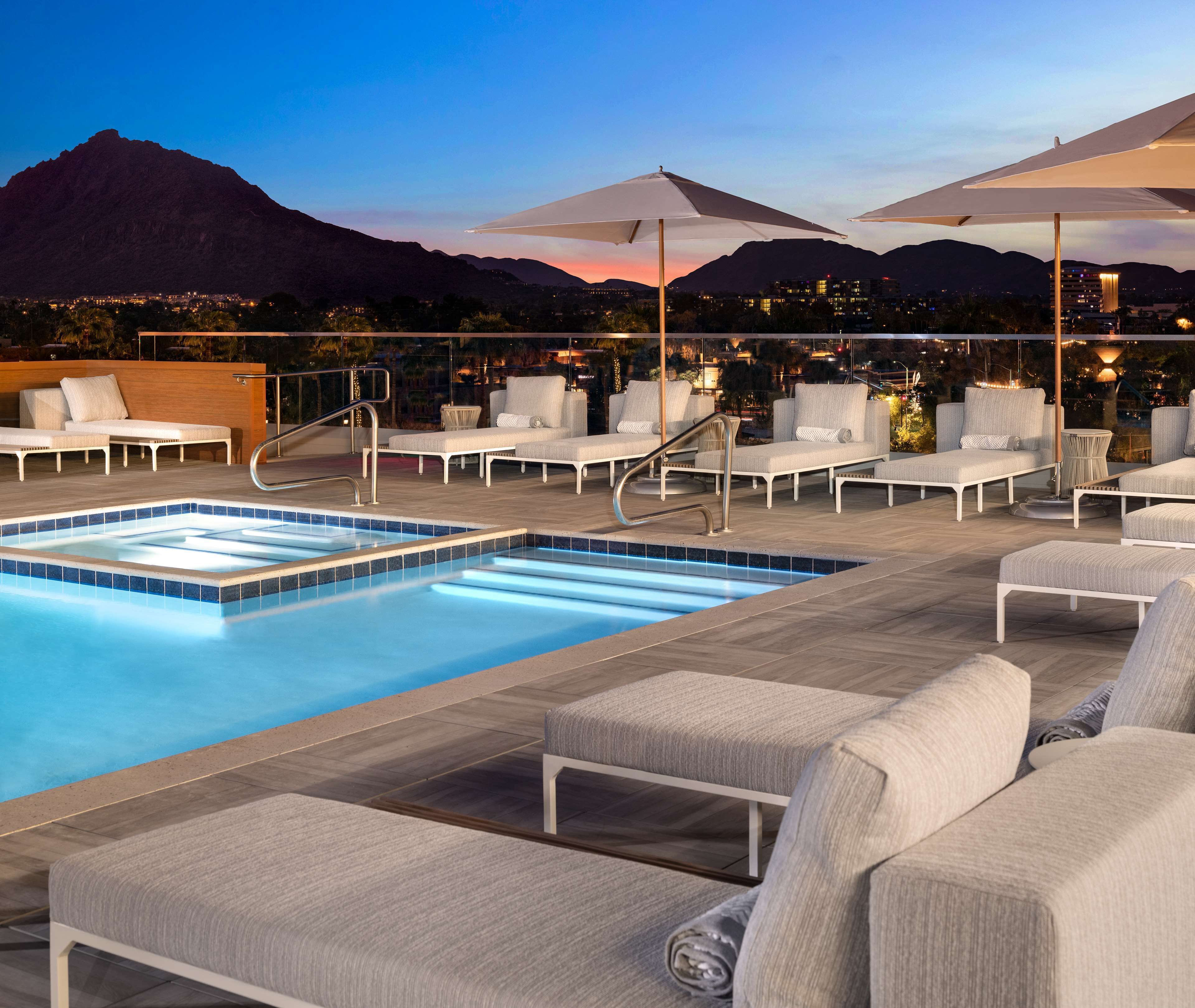A rooftop pool area with lounge chairs and umbrellas, overlooking a cityscape and mountains.
