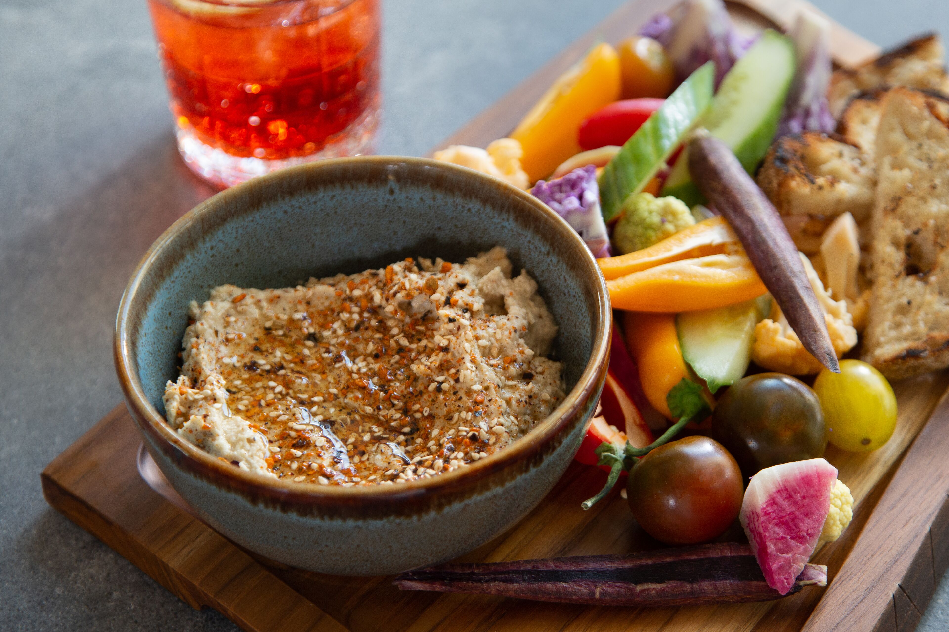 Un tazón de hummus acompañado de varios vegetales y pan sobre una tabla de madera.