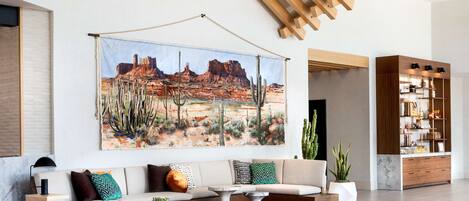 A modern living room with a large painting of a desert landscape, a sectional sofa with various cushions, a coffee table, and a wooden cabinet with shelves.