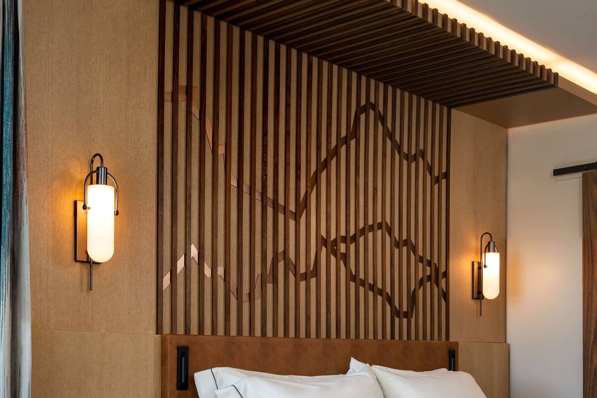 Una habitación de hotel moderna con cabecera de madera, dos lámparas de noche, una mesita de noche con un teléfono y libros, y una cama con sábanas blancas.
