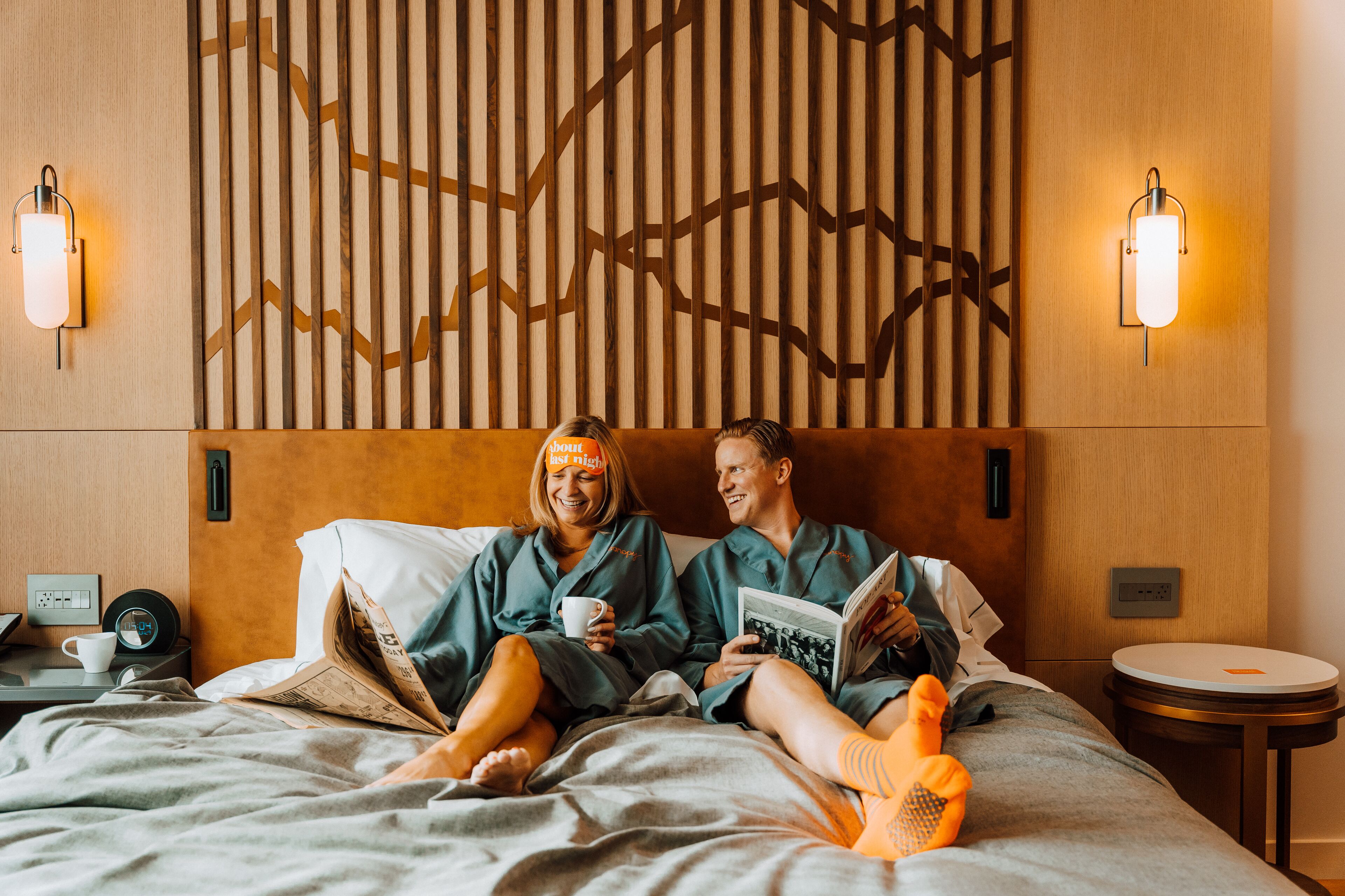 Um casal de pijamas iguais em uma cama com cabeceira de madeira, lendo um livro e segurando uma caneca.